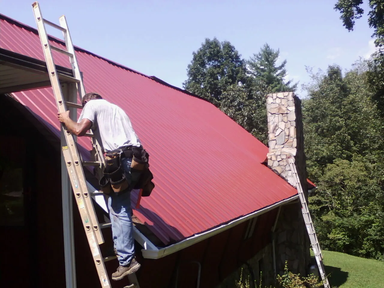 Roofer on a ladder beside a red metal roof and stone chimney — Soffit & Fascia Repair by Roofing Brothers in Upper Falls