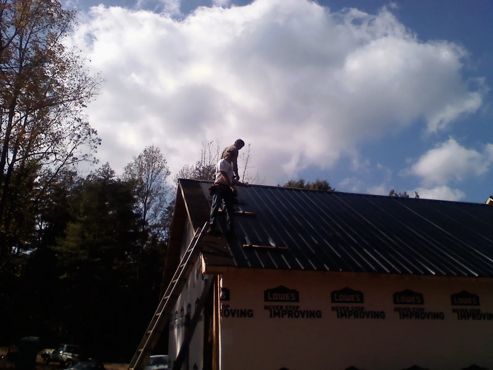 Roofer fastening dark metal panels high against a cloudy sky, from a Roof Inspection job in Upper Falls